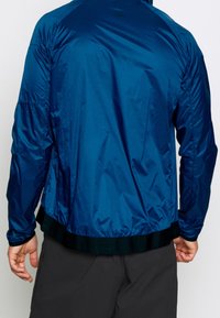 Man i en glänsande blå jacka med elastisk fåll och svarta byxor, sedd bakifrån mot en enkel bakgrund.