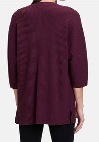 Žena v tmavě fialovém svetru s dlouhým rukávem a žebrovaným vzorem, s postranními rozparky, zobrazená zezadu na jednobarevném pozadí.