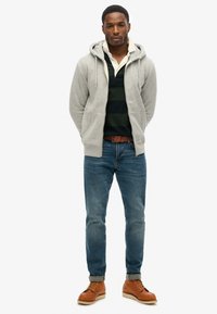 Felpa grigia con cappuccio sopra una polo a strisce verde scuro, jeans di denim blu e stivali brown con lacci. Outfit semplice e casual.