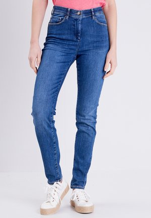 Hoch geschnittene blaue Jeans aus Denim mit einer taillierten Passform, fünf Taschen und sichtbaren Nähten. Kombiniert mit weißen Sneakers mit Jutesohlen.