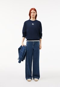 Felpa navy con logo bianco Lacoste, abbinata a jeans a gamba larga blu. Il modello tiene una giacca di jeans. Tessuto morbido, vestibilità casual.