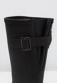 Botas de lluvia de goma negras con un borde superior redondeado, detalles decorativos de correa y hebilla. Textura suave con un acabado brillante.
