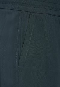 Zbliżenie na ciemną tkaninę z przeszytą skośną kieszenią i detalem elastycznego pasa.