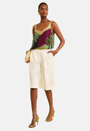 Nő zöld-lila leveles mintájú ujjatlan felsőben arany szegéllyel, fehér térdig érő rövidnadrágban, arany retiküllel és olívazöld magassarkúban.