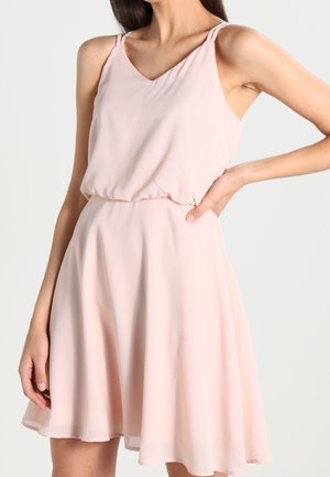 Kobieta ubrana w bezrękawnikową, jasnoróżową sukienkę do kolan z cienkimi podwójnymi ramiączkami, dekoltem w serek i marszczonym taliowaniem, stojąca z jedną ręką na biodrze.