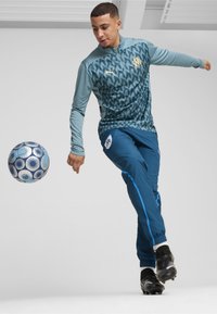 Šviesiai mėlyna sportinė striukė su geometriniu raštu, derinama su tamsiai mėlynomis kelnių, turinčiomis šonines juostas. Juodi sportbačiai ir mėlynas raštuotas futbolo kamuolys.