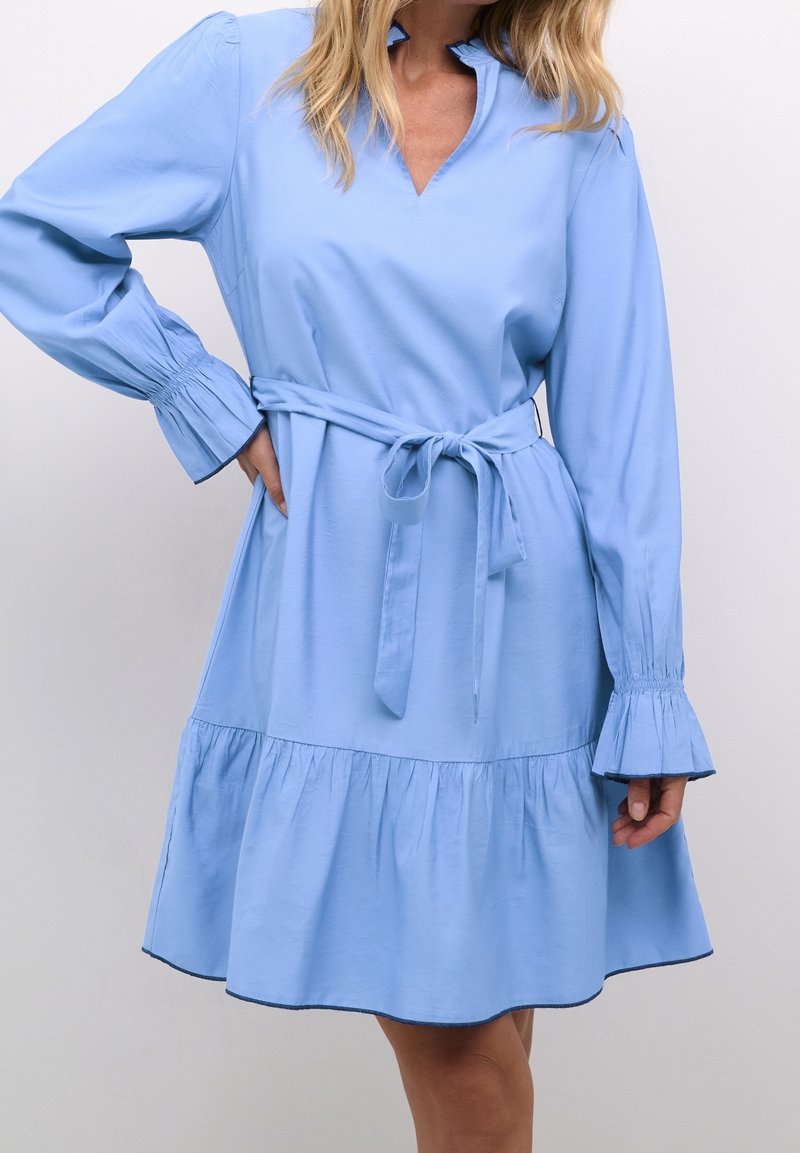 Une femme porte une robe bleu clair à manches longues avec des poignets à volants et une ceinture nouée à la taille, sur un fond uni.
