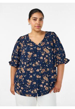 Mujer que lleva una blusa azul marino con estampado floral y mangas abullonadas, y pantalones blancos, de pie frente a un fondo blanco liso.