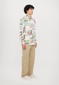 Valge särk, millel on värvilised rannateemalised mustrid, nööpidega ees, koos beežide pükste ja valgete spordijalatsitega. Sile kangas, pikad varrukad.