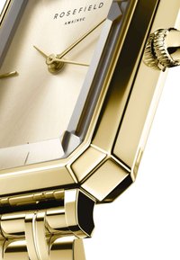 Reloj rectangular tono dorado con una esfera dorada texturizada, manecillas delgadas y una correa de metal pulido con un diseño angular y una corona.