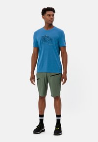 Heren blauwe overhemd met korte mouwen en bergafbeelding, gecombineerd met groene shorts met zwarte ritszakken. Zwarte sneakers en gestreepte sokken.