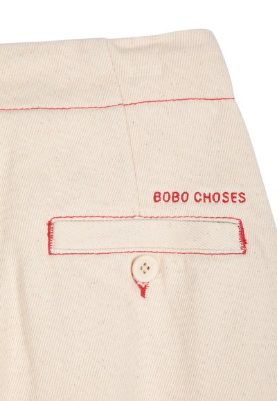 Tissu beige avec une poche boutonnée soulignée par une couture rouge et le nom de la marque "BOBO CHOSES" brodé au-dessus en fil rouge.