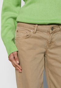 Calças de algodão caqui com um design justo, apresentando rebites de cor cobre, múltiplos bolsos e uma cintura de altura média.