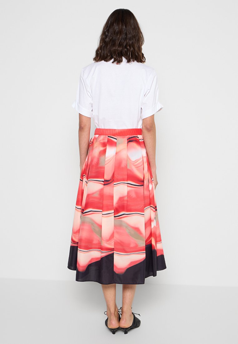 Kobieta stoi tyłem, ubrana w białą koszulę z krótkim rękawem i plisowaną spódnicę midi w abstrakcyjne wzory w odcieniach czerwieni, różu, czerni i beżu.