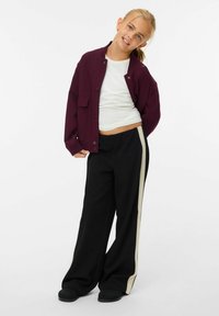 Giacca bordeaux con bottoni a pressione sopra una maglia bianca, abbinata a pantaloni larghi neri con una striscia bianca lungo il lato. Tessuto morbido e vestibilità rilassata.