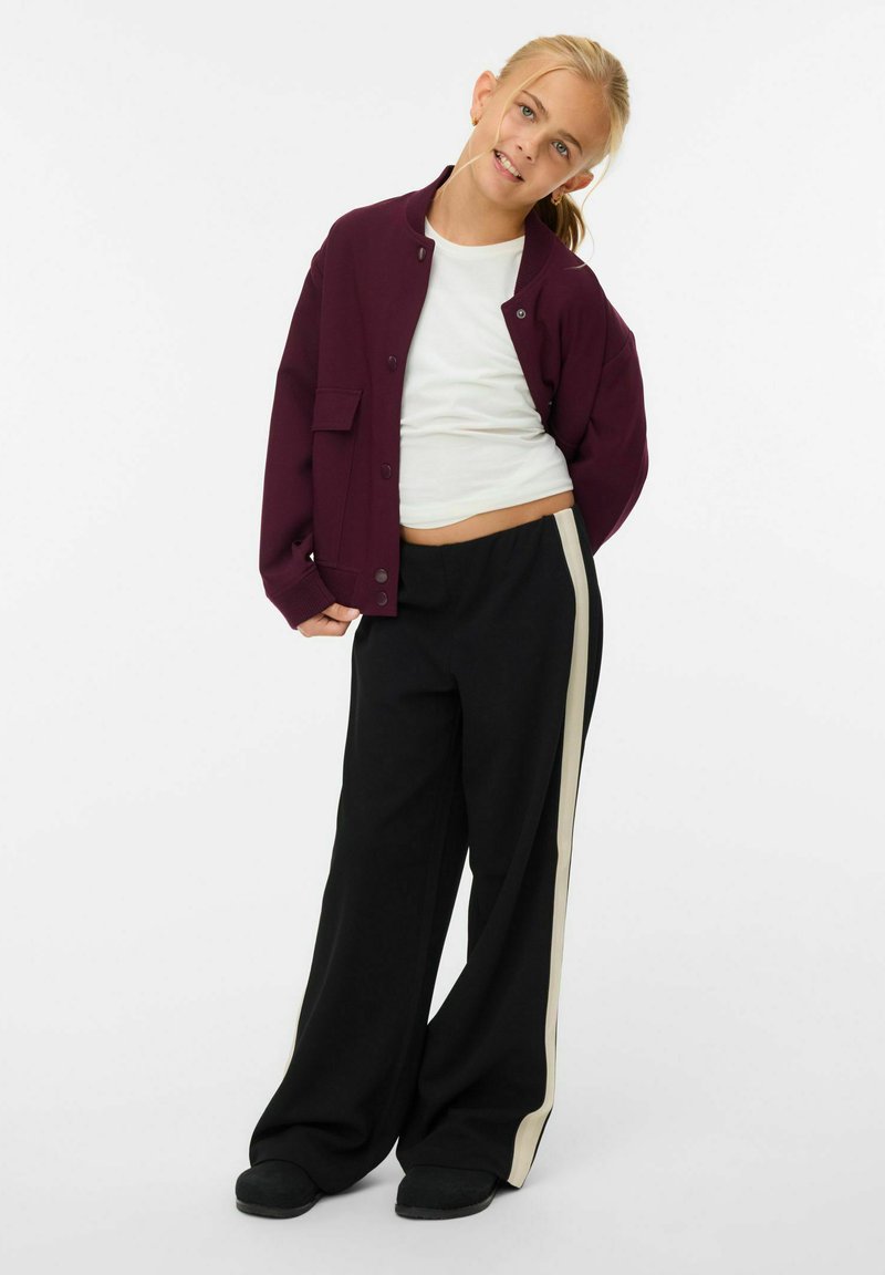 Giacca bordeaux con bottoni a pressione sopra una maglia bianca, abbinata a pantaloni larghi neri con una striscia bianca lungo il lato. Tessuto morbido e vestibilità rilassata.