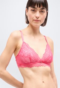 Νεαρή γυναίκα που φοράει ένα ροζ μπραλέτο με λεπτές τιράντες από δαντέλα, στέκεται μπροστά σε απλό ανοιχτό γκρι φόντο.