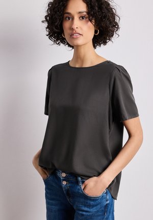 Frau mit lockigem Haar, die goldene Creolen trägt, eine schwarze Kurzarmbluse und blaue Jeans, mit den Händen in den Taschen, vor einem einfarbigen Hintergrund stehend.