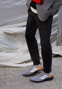 Persona che indossa sneaker Adidas grigie e arancioni con strisce riflettenti, pantaloni jogger neri, in piedi su ghiaia vicino a un telone bianco.