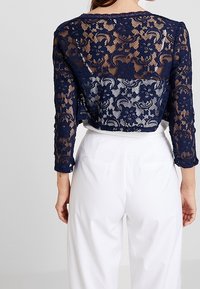 Marinblå spets crop top med intrikat blommönster och långa ärmar, i kombination med vita högmidjade byxor med bakfickor.