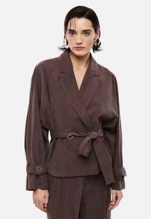 Donna con capelli corti e scuri, che indossa grandi orecchini a cerchio dorati e una giacca di suede marrone con cintura e pantaloni abbinati, su uno sfondo semplice.