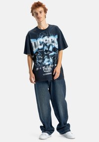 Mladý človek so krátkymi červenými vlasmi nosí nadrozmerné tmavé tričko s potlačou, voľné modré džínsy a biele tenisky, stojí pred jednoduchým pozadím.