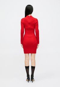 Rdeča rebrasta pletena obleka z dolgimi rokavi, visokim ovratnikom, prilegajočim silhueto in zadrgo na hrbtni strani. Ujemajoče se z črnimi nogavicami in koničastimi petami.