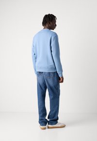 Γαλάζια φούτερ με στρογγυλή λαιμόκοψη, ριμπ μανίκια και στρίφωμα. Φοριέται με χαλαρά μπλε τζιν και λευκά αθλητικά παπούτσια. Ουδέτερο φόντο.