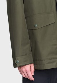 Olivovo zelená bunda vyrobená z hladkej látky, s gombíkovým manžetovým uzáverom a náprsním vreckom so preklápacím klopom.