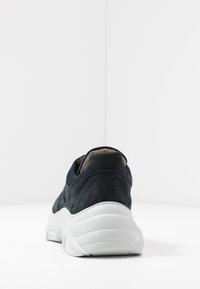 Sneaker blu navy con parte superiore in suede e mesh, vista dal tallone, caratterizzata da una spessa suola bianca testurizzata su uno sfondo bianco.