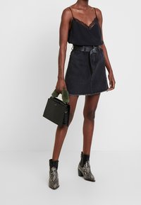 Kvinna i svart camisole med spetskant, svart denimkjol med bälte, snakeskinnsmönstrade ankelstövlar, håller en svart handväska med handtag i grön päls.