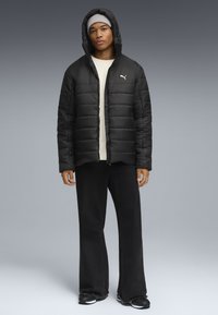 Giubbotto imbottito nero con chiusura a zip e cappuccio, con logo bianco. Il modello indossa pantaloni neri a gamba larga e scarpe sportive.