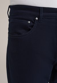 Tamsiai mėlyni džinsai su lygiu paviršiumi, turintys priekinį kišenę, sidabrinius varžtus ir išsamią siuvinėjimo detalę palei siūles.