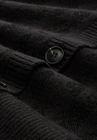 Zwarte gebreide cardigan met geribbelde textuur en subtiele polkadotpatronen, voorzien van een ronde zwarte knoopsluiting.