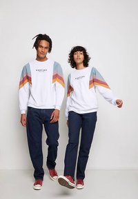 Balti džemperi ar krāsainām četrstūra svītrām persiku, zilā un sinepju krāsā, kombinēti ar tumšām džinsu biksēm un sarkaniem sporta apaviem. Krekliem ir melns logotipa teksts.