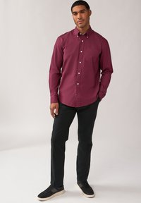 Camicia a maniche lunghe color bordeaux con colletto, taschino sul petto e bottoni bianchi, abbinata a pantaloni neri e sneaker scure.