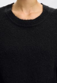 Prim-plan al gâtului și părții superioare a pieptului unei persoane care poartă un pulover negru din lână tricotată pe un fundal deschis.