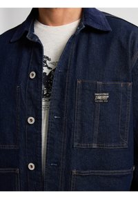 Mørkeblå denimjakke med en kontrasterende krave, to forlommer, metalknapper og synlige syninger. Bæres over en lys grå skjorte.