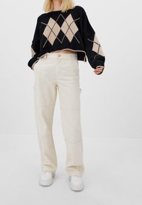 Mujer vistiendo pantalones anchos beige, zapatos gruesos blancos y un suéter corto negro con un patrón de rombos beige.