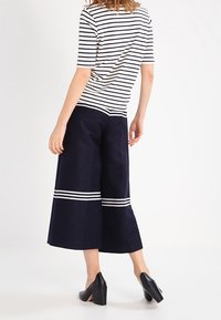 Žena v bílém a černém pruhovaném topu s námořnickými širokými kalhotami ke kolenům, které mají bílé horizontální pruhy, a černých blokových podpatcích.