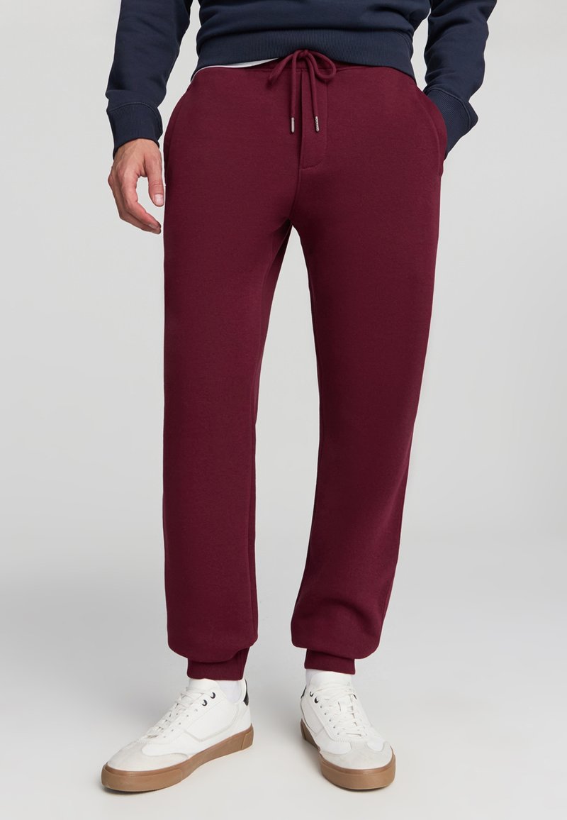 Pantaloni della tuta bordeaux con vita regolabile tramite cordoncino, polsini a costine, tasche laterali e una texture morbida, abbinati a sneakers bianche e calzini grigi.