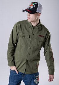 Camicia in cotone verde con due tasche sul petto e chiusura con bottoni; presenta dettagli ricamati e motivi decorativi sulle maniche e sull'orlo.