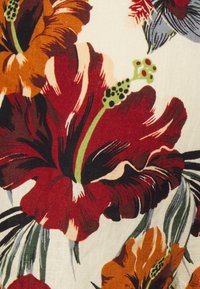 Květinová látka s velkými, živými květy hibiscus v červené a oranžové barvě, s zelenými akcenty, na krémovém pozadí.