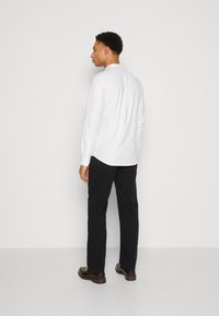 Camisa blanca de manga larga con dobladillo curvado, combinada con pantalones oscuros y zapatos marrones. La camisa tiene un detalle de botones en los puños.