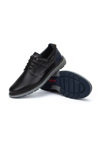 Scarpe in pelle nera con finitura liscia, collo imbottito e suola in gomma grigia. Presentano lacci e dettagli di cucitura sottili. Visibile etichetta del marchio.