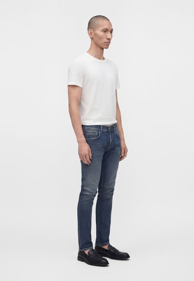Homme debout portant un t-shirt blanc uni, un jean bleu ajusté et des mocassins en cuir noir sur un fond blanc uni.
