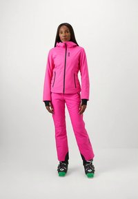 Casaco de esqui cor-de-rosa brilhante com capuz e fecho de correr, combinado com calças de esqui cor-de-rosa correspondentes. Apresenta detalhes a preto e botas de esqui robustas.