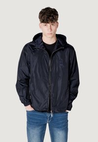 Giacca a vento blu navy con chiusura zip, cappuccio con cordino e polsini elastici. Indossata sopra una maglietta nera e abbinata a jeans blu.