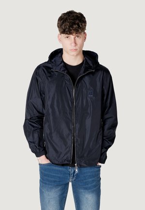 Giacca a vento blu navy con chiusura zip, cappuccio con cordino e polsini elastici. Indossata sopra una maglietta nera e abbinata a jeans blu.