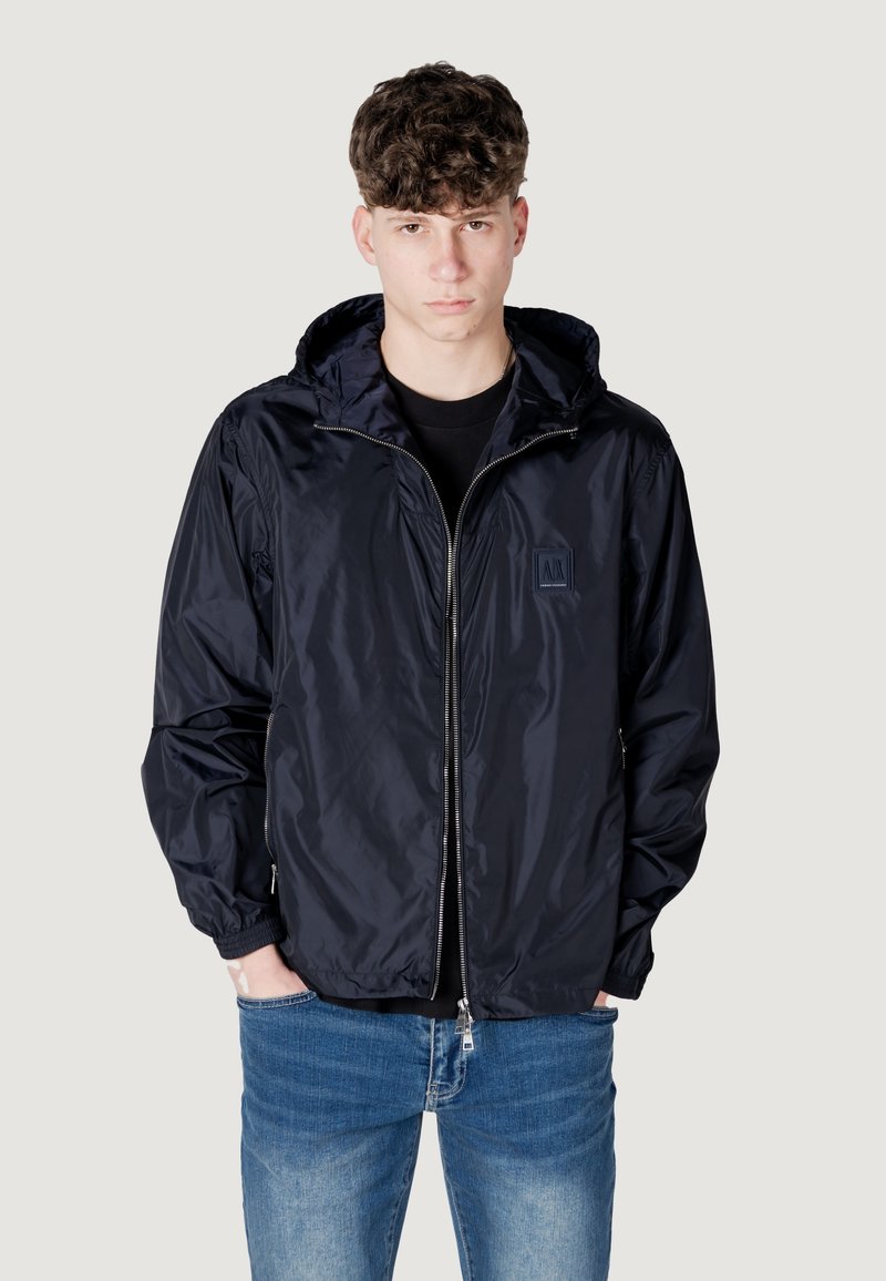 Giacca a vento blu navy con chiusura zip, cappuccio con cordino e polsini elastici. Indossata sopra una maglietta nera e abbinata a jeans blu.